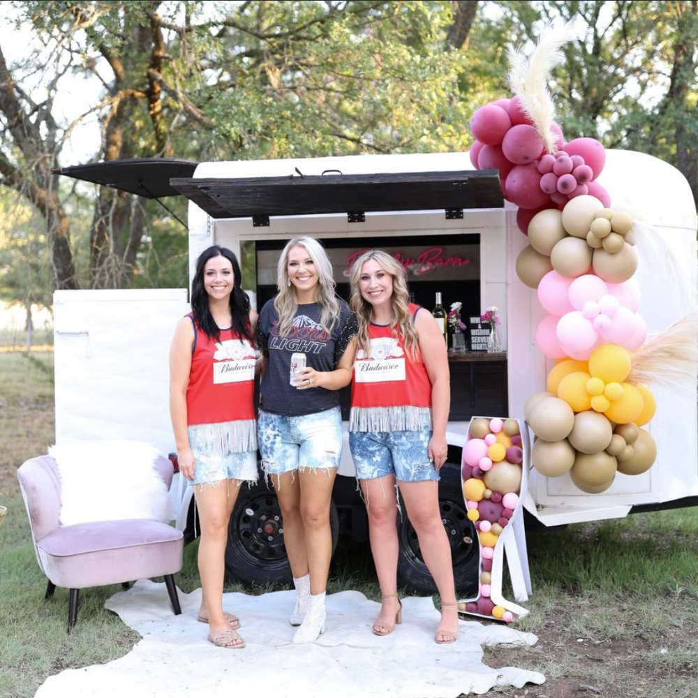 Ladies celebrate a bridal shower and smile for the camera in front of a mobile bar trailer in Austin.